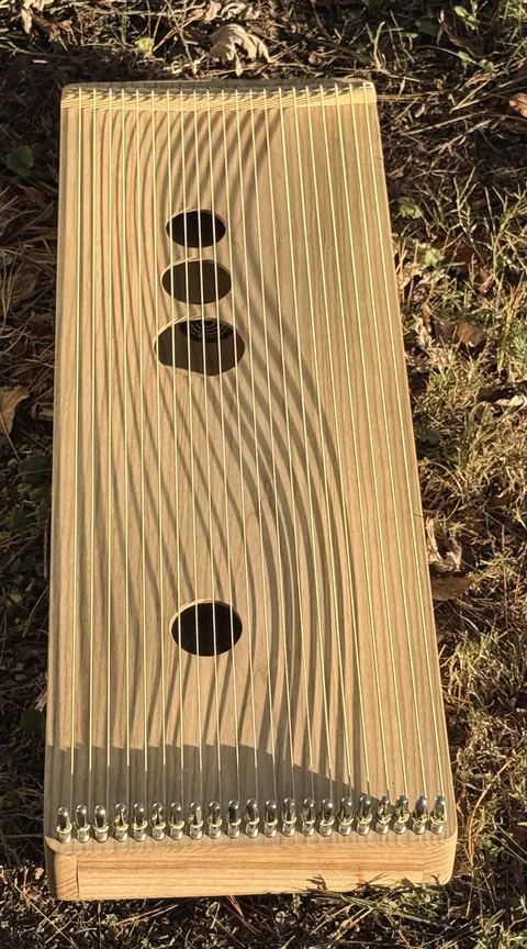 A wooden monochord with wavy grain lying on the grass, its many strings catching the light